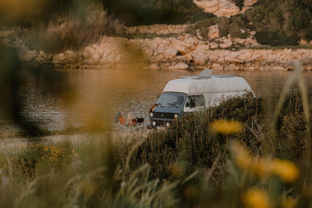 VW T3 Postbus steht in der Nähe vom Meer, im Vordergrund befinden sich Pflanzen.