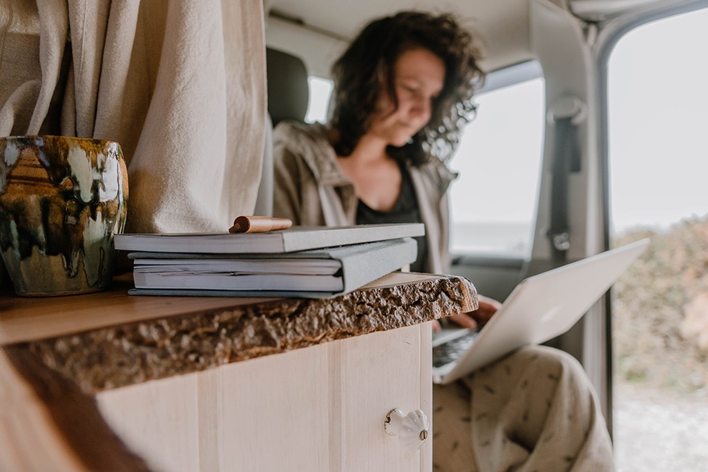 Person sitz auf gedrehter Doppelsitzbank in VW Bus und arbeitet, im Vordergrund liegen Notitzbücher.