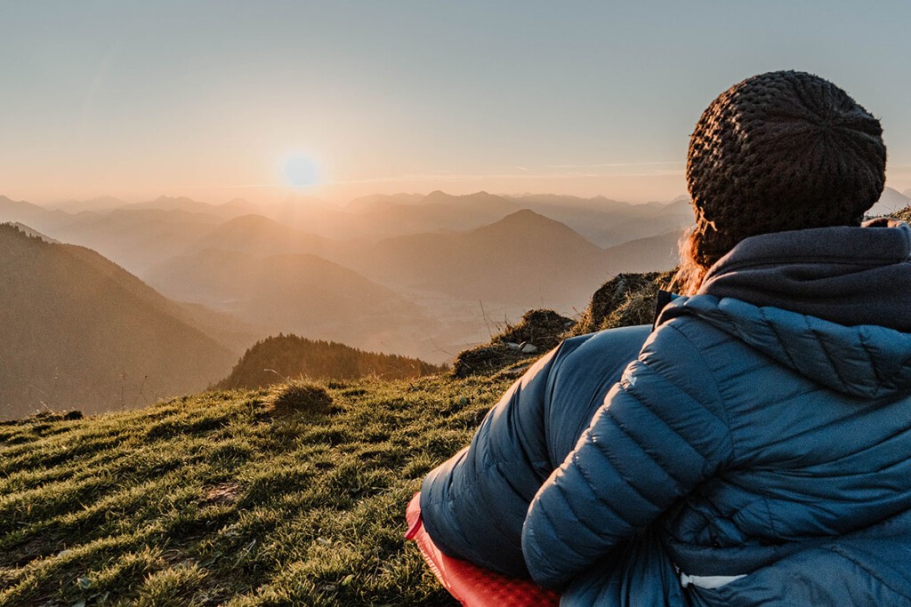 Inspiration für Camping Geschenke: Schlafsack. Person sitzt im Schlafsack auf Isomatte auf Berggipfel und beobachtet Sonnenaufgang.