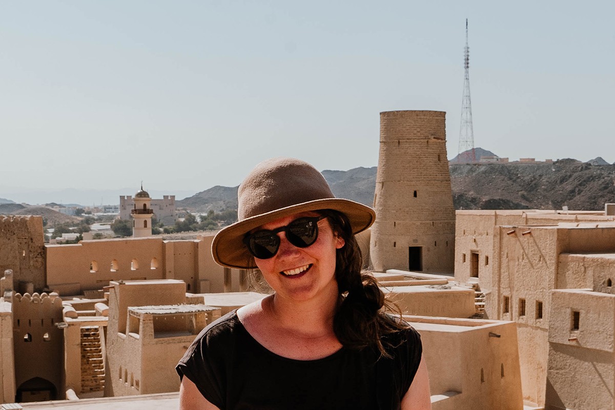 Elisa Model vom Camping Blog take an adVANture trägt Hut, schwarze Sonnenbrille, schwarzes Kleid und blickt in die Kamera. Im Hintergrund befindet sich eine alte Siedlung aus Lehm.