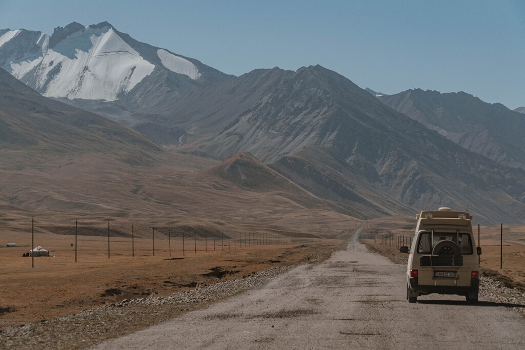 VW T4 Camper fährt auf schmalem und löchrigen Pamir-Highway auf der kirgisischen Seite vor der Grenze. Schneebedeckte Berge befinden sich im Hintergrund.
