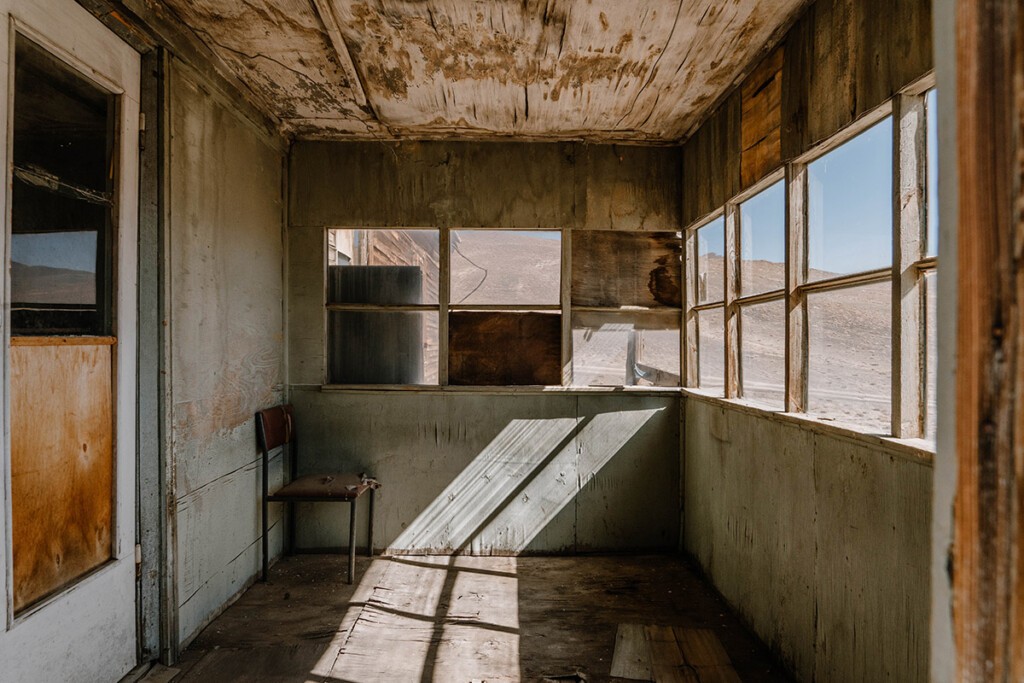 Blick in den hölzernen Vorraum von einem verlassenen Gebäude. Durch die Fenster scheint die Sonne, in einer Ecke steht ein Stuhl.