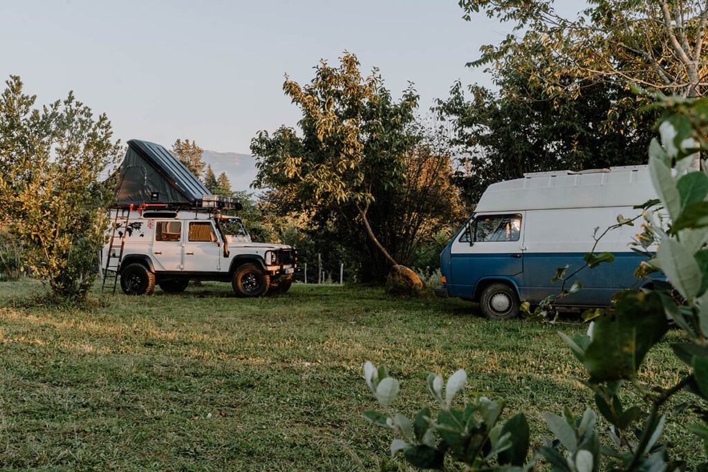 Camping Georgien auf Campingplatz: Blau-weißer VW T3 mit Hochdach und weißer Landrover Defender mit aufgeklapptem Dachzelt stehen auf grüner und von Bäumen umgebener Wiese vom Camp Itara.