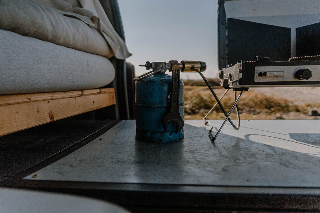 Kleine, blaue und wiederbefüllbare Gasflasche steht auf Heckauszug im Camper und ist an Campingkocher angeschlossen.