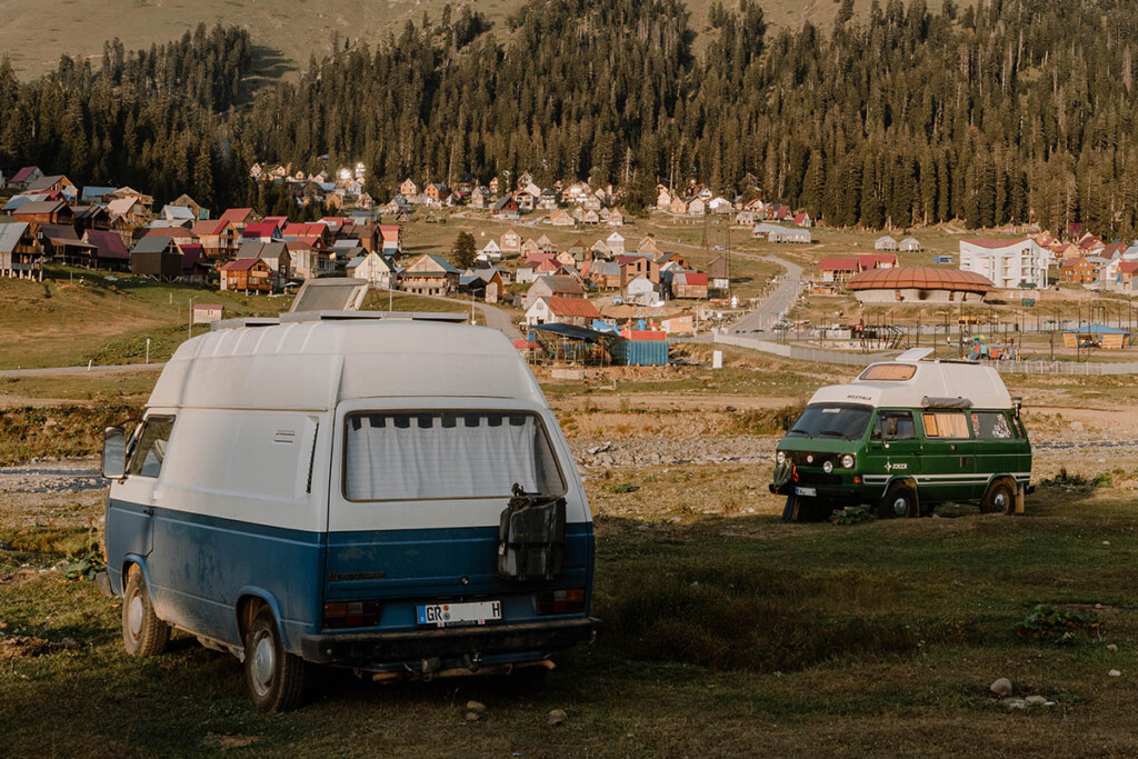 Wildcampen in Georgien: Zwei VW T3, einer grün mit weißem Hochdach, einer blau-weiß mit Hochdach, stehen auf einer Wiese in den Bergen, im Hintergrund liegt ein Ort am Berghang.