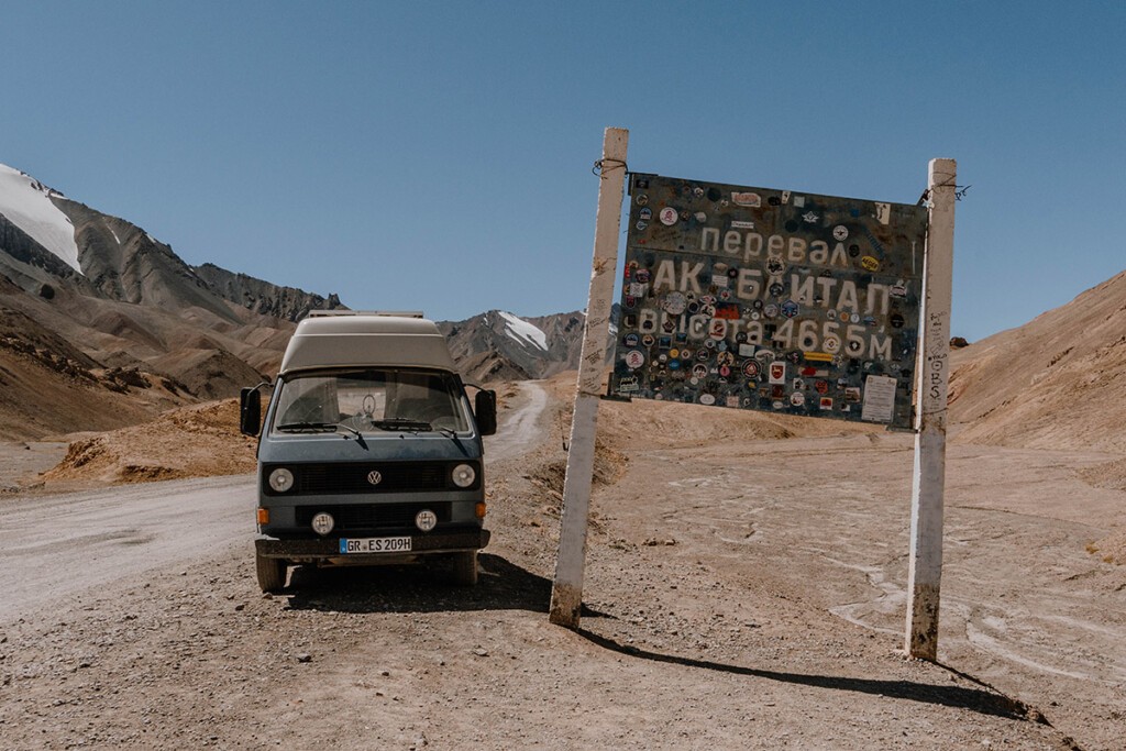 Blau-weißer VW T3 Camper steht neben dem mit Stickern überklebten Schild vom Ak-Baital-Pass am Pamir-Highway. 