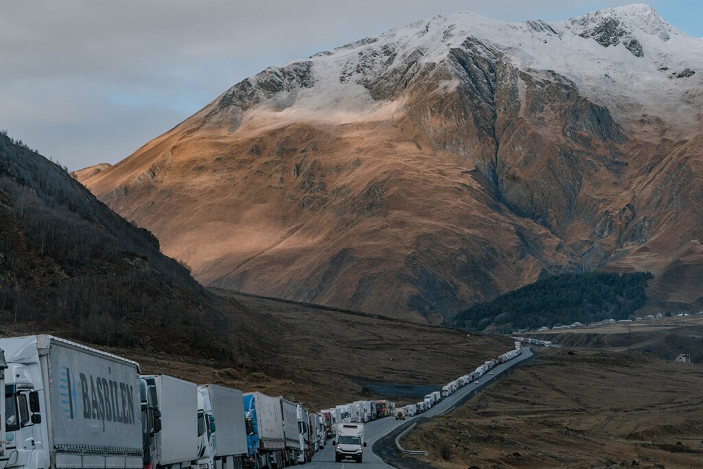 Lange LKW-Schlange am Straßenrand, herbstliche Berge mit Schnee im Hintergrund.