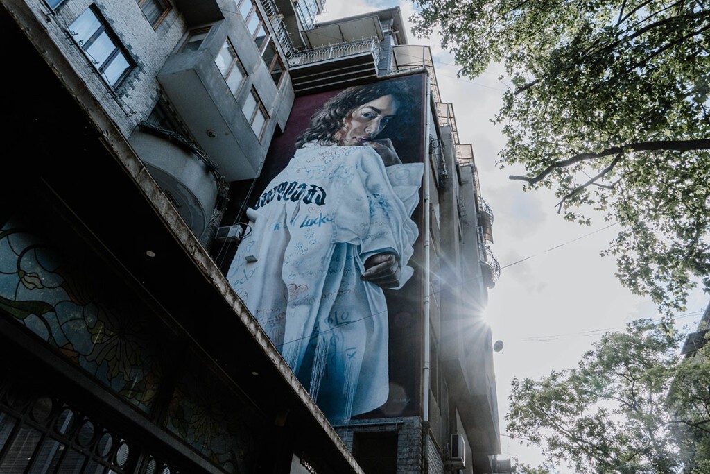 Street-Art an Hauswand in Tbilisi. Abgebildet ist eine Frau in weißer Jacke mit Schriftzügen drauf.