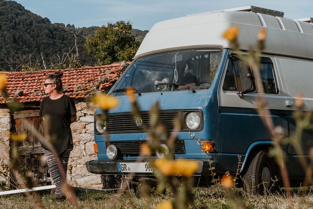 Elisa Model vom Vanlife Blog take an adVANture steht neben ihrem VW T3 Camper mit Hochdach und vor einem kleinen Steinhaus in Italien auf einer Wiese.