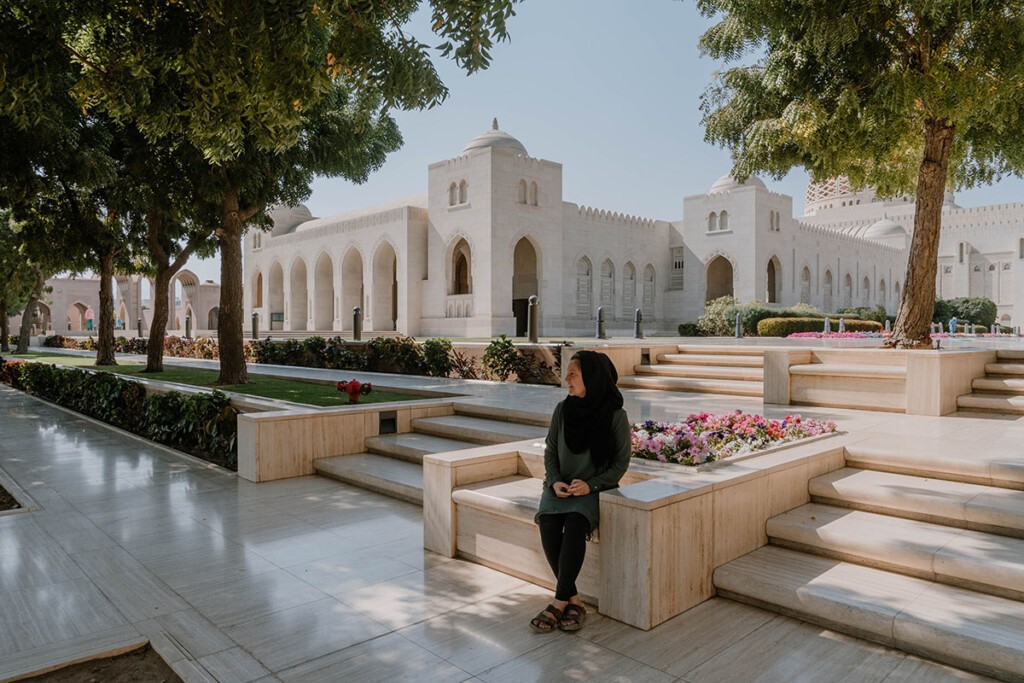 Frau sitzt auf Mauer aus Marmor vor Moschee im Oman, Bäume spenden Schatten.