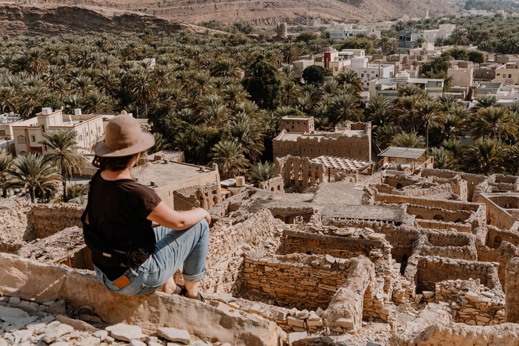 Elisa Model vom Camping Blog take an adVANture sitzt auf Wand über einer alten, verfallenen Lehmstadt im Oman. Sie trägt einen Hut, ein schwarzes Oberteil und Jeans. Im Hintergrund sieht man Palmen.
