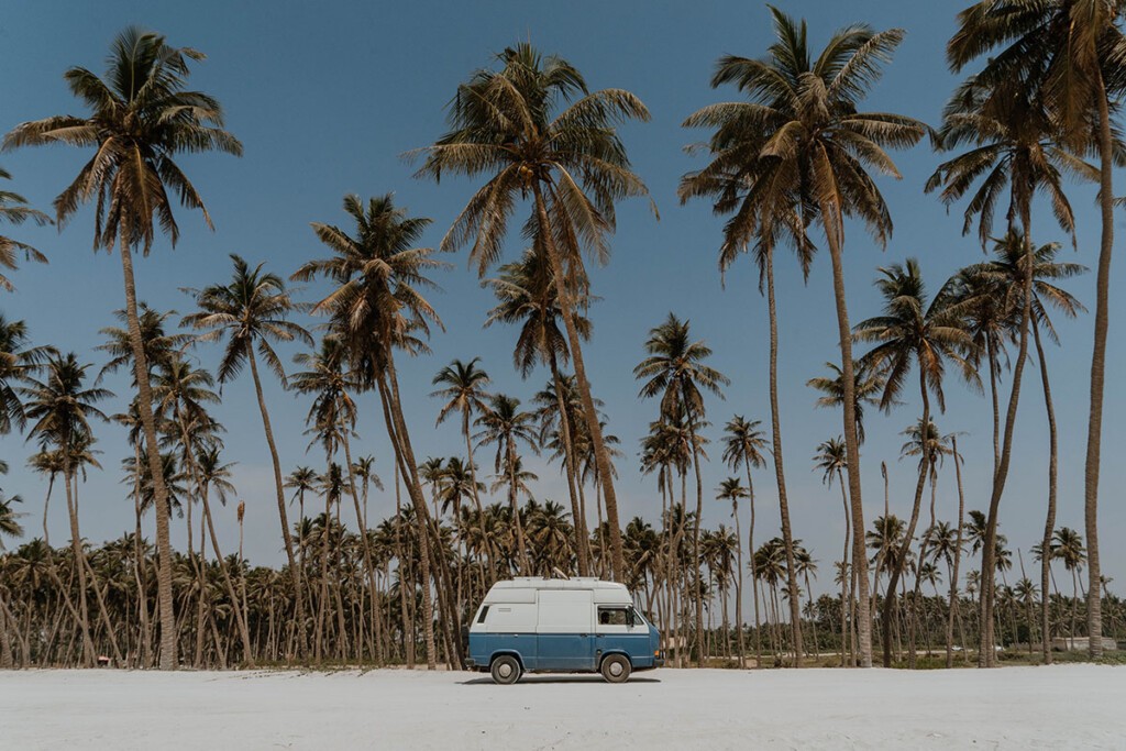 Blau-weißer VW T3 Camper steht auf weißem Strand mit Palmen im Hintergrund.