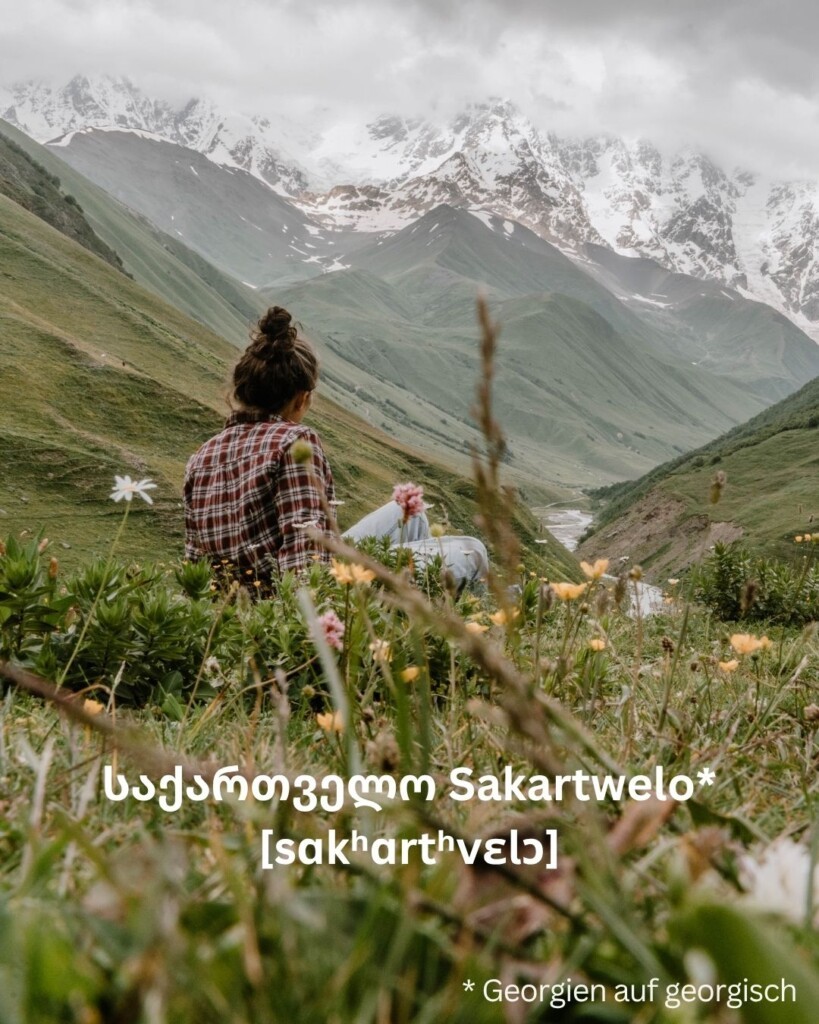 Frau sitzt auf Wiese und blickt in Richtung Schchara, dem höchsten Berg in Georgien. Davor erstreckt sich ein Tal.