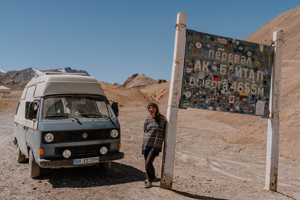 Elisa Model vom Camping Blog take an adVANture lehnt am Schild vom Ak-Baital-Pass in Tadschikistan, daneben steht ihr VW T3.