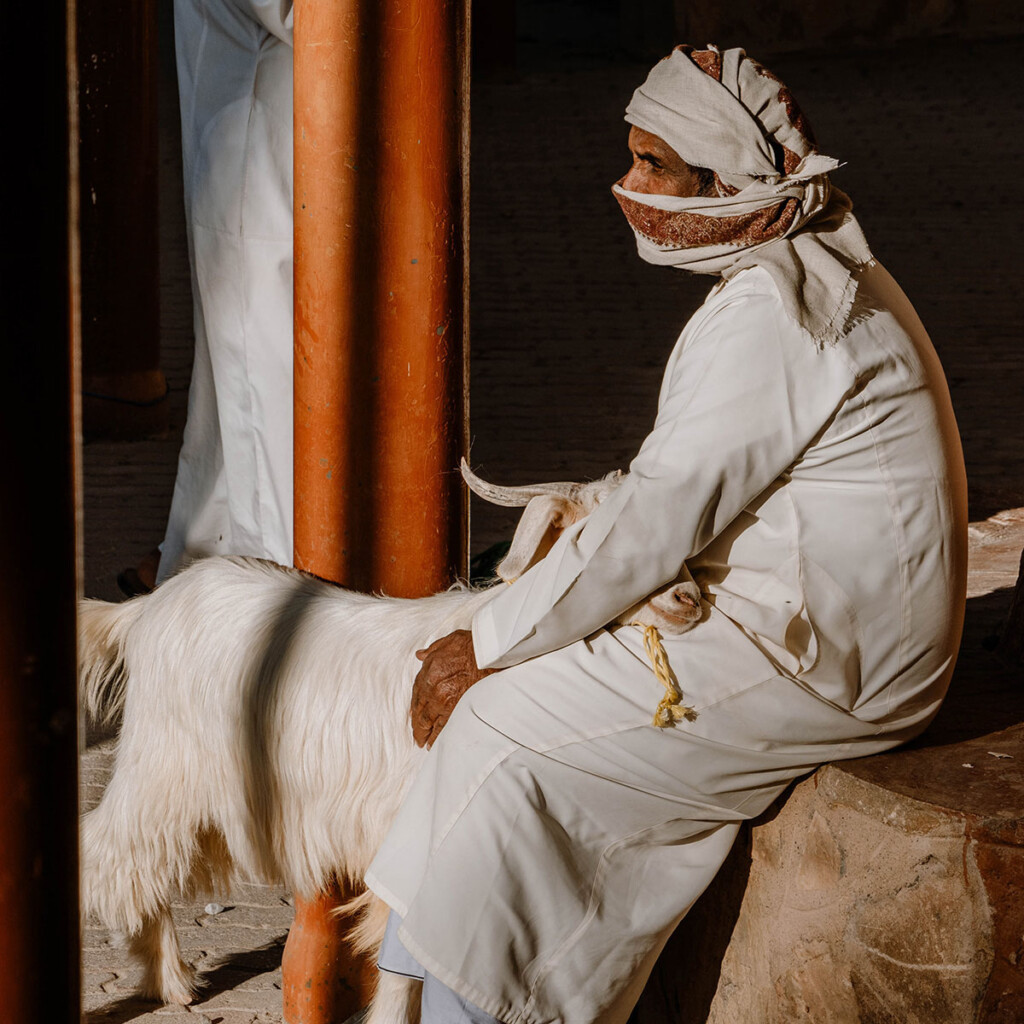 Omani sitzt in traditioneller Kleidung auf Mauer und streichelt Ziege.