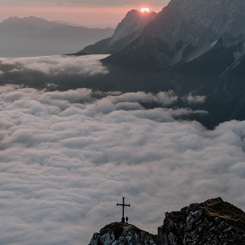 Sonnenaufgang mit Blick vom Grubigstein auf Wolkenmeer und Gipfelkreuz mit Mensch